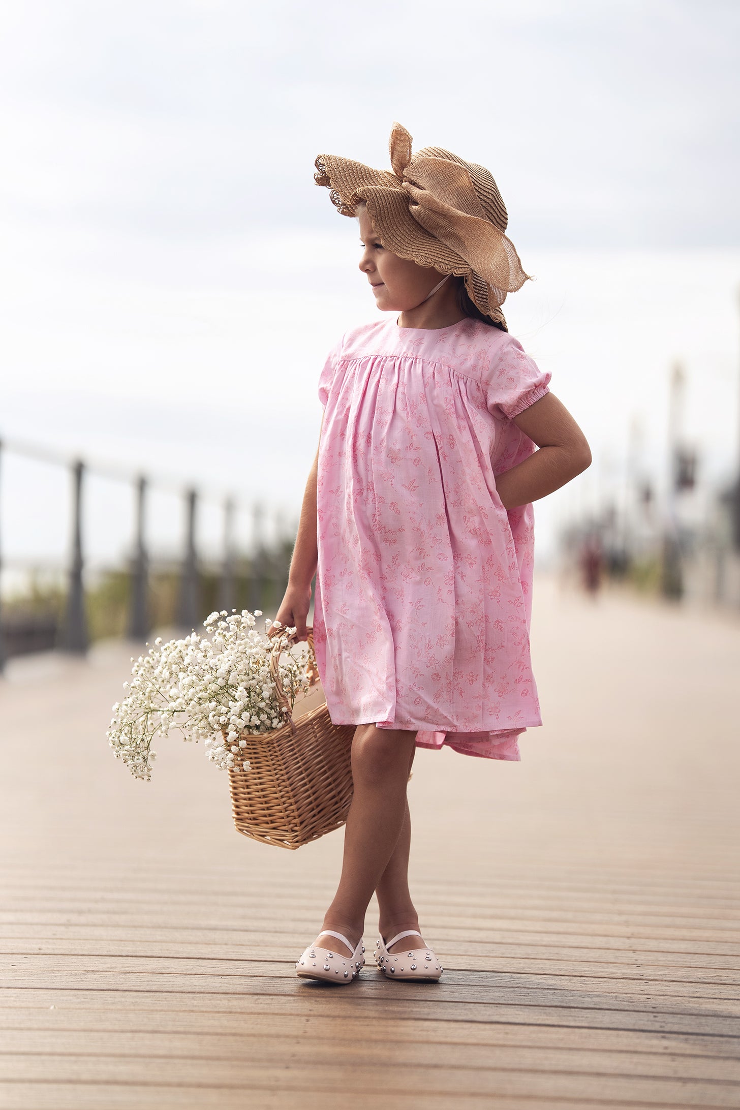 Young girl in pink flower dress with short sleeves, straw hat, carrying a wicker basket of white flowers on boardwalk.