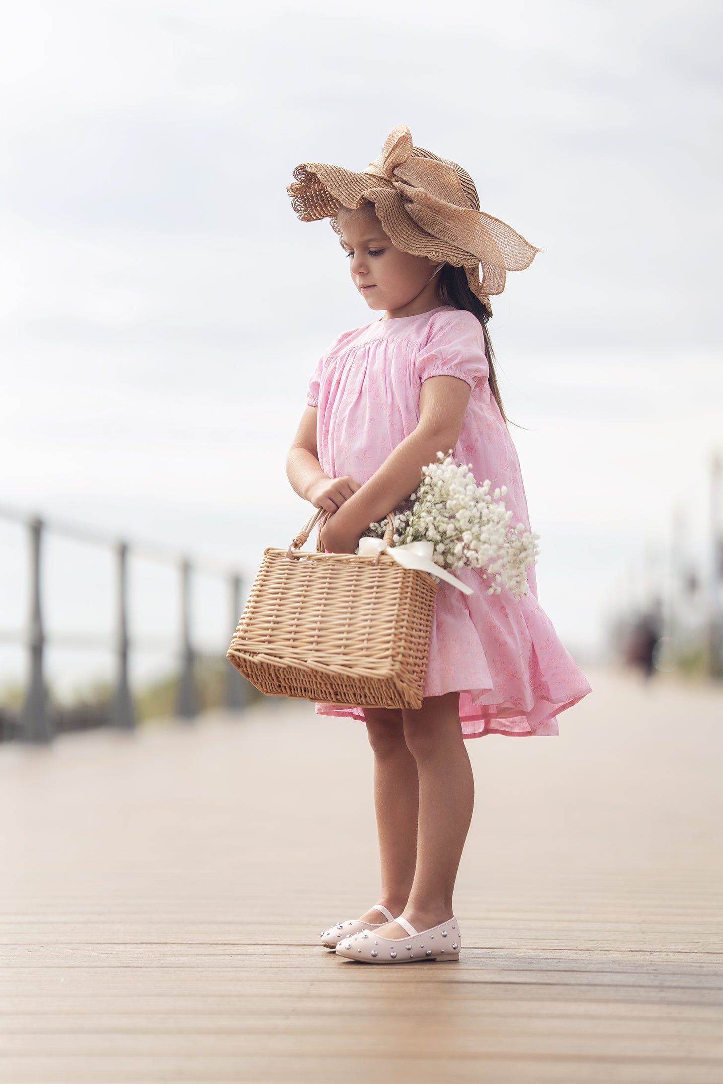 Girl in pink short-sleeve dress with wide-brim straw hat, carrying a woven basket of white flowers.