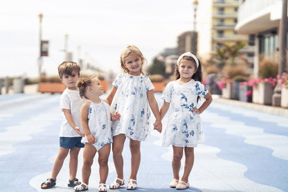 Children in white and blue floral outfits inspired by Blue Flower Skirt, walking hand in hand on a sunny boardwalk.