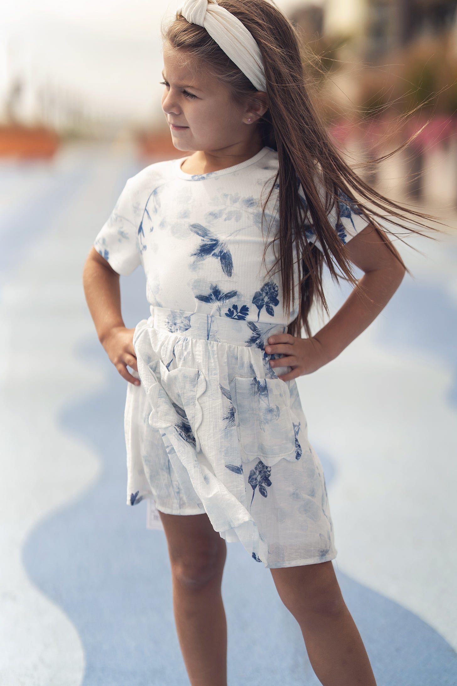 Girl wearing a blue flower print short-sleeve top with a light skirt, outdoors with a headband.