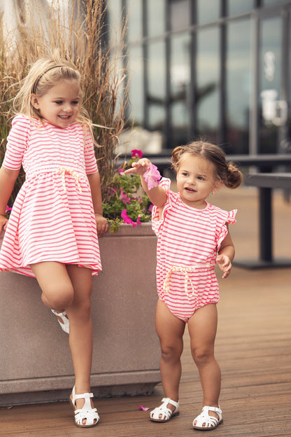 Two young girls in neon pink striped dresses outdoors, one with 3/4 sleeves and white sandals.