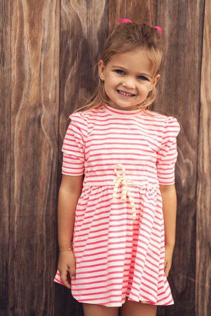Smiling girl wearing neon pink and white striped dress with a yellow waist bow against a wooden backdrop.