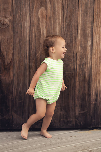 Toddler wearing neon green striped romper with pockets, smiling on a wooden deck.