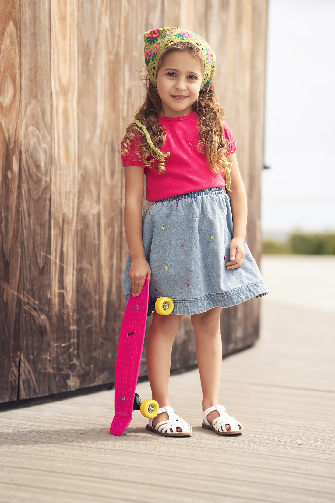 Girl wearing Embroidered Heart Denim Skirt with pink top and white sandals, holding a pink skateboard.