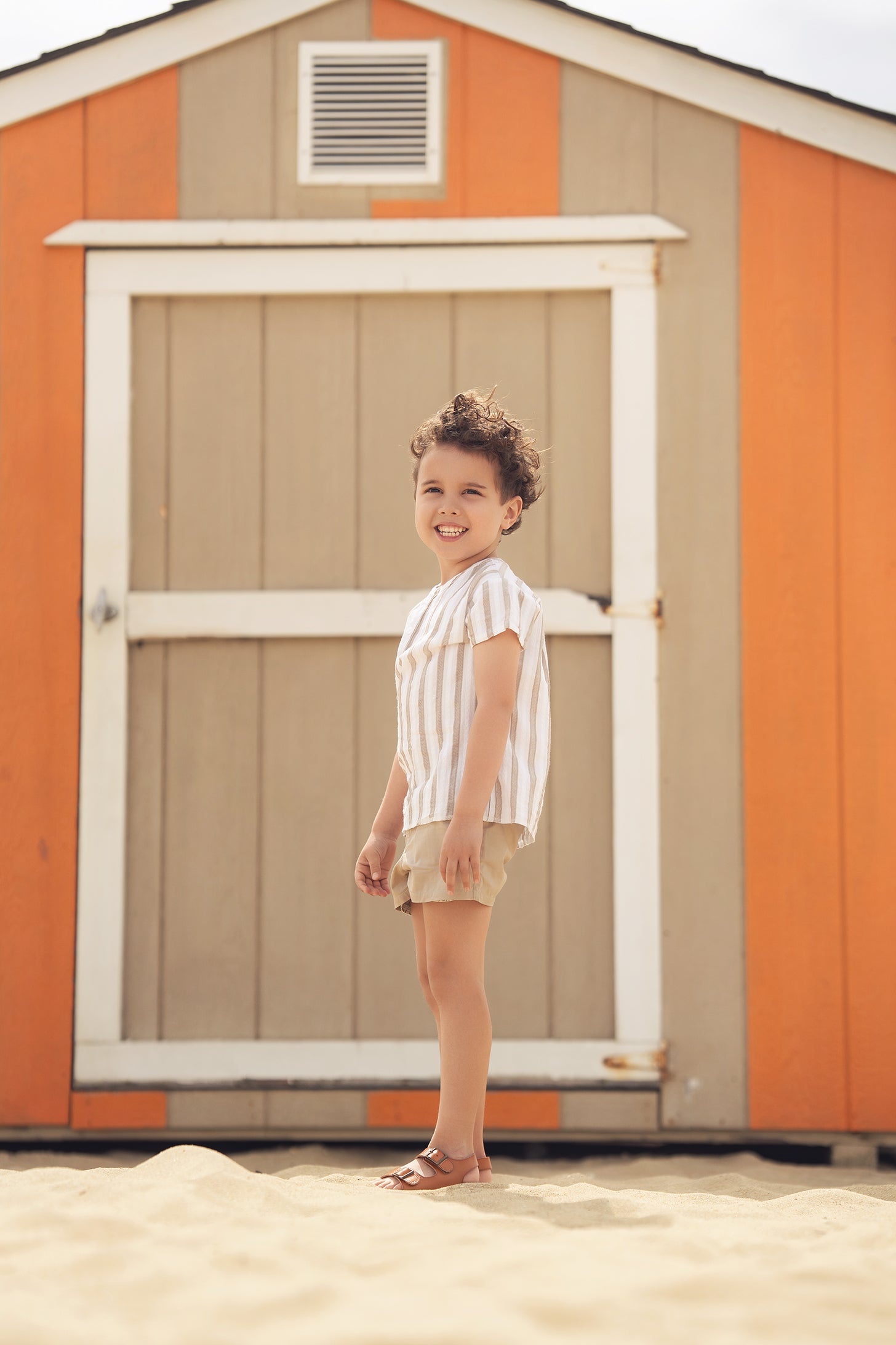 Tan and White Striped Shirt worn by a smiling child at the beach in front of an orange shed.