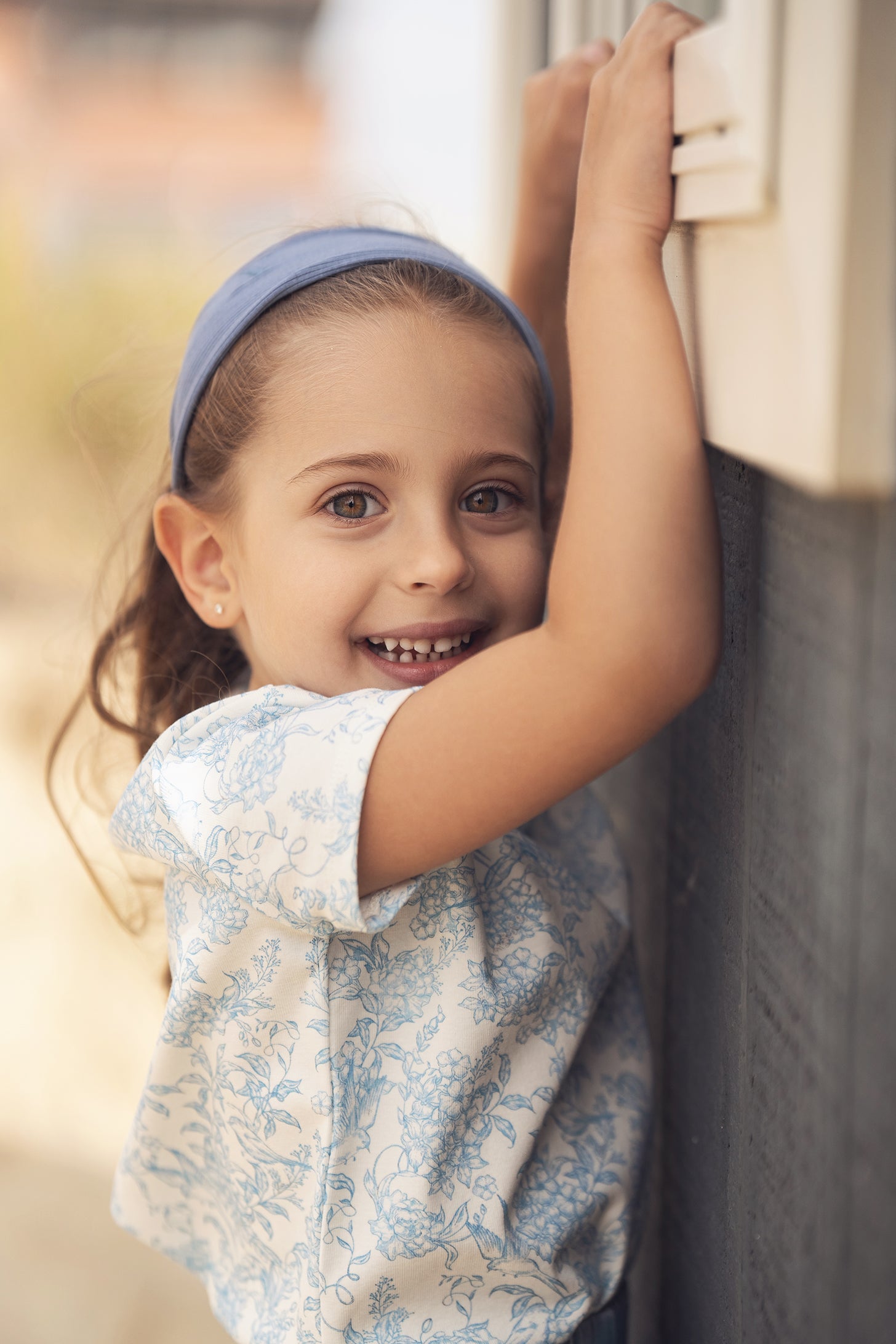 Girl wearing blue flowered short-sleeve T-shirt, smiling, with a blue headband, leaning against a wall.