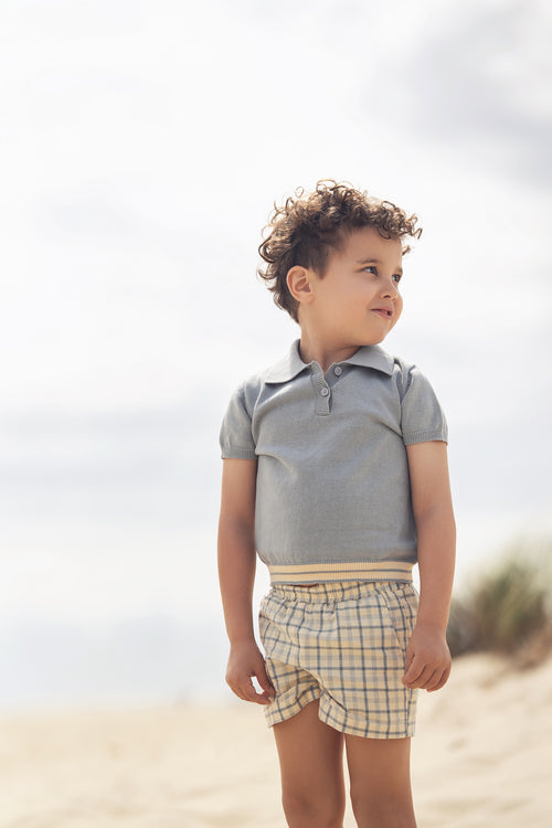 Young boy wearing a light blue knit sweater and plaid shorts on a sunny beach.