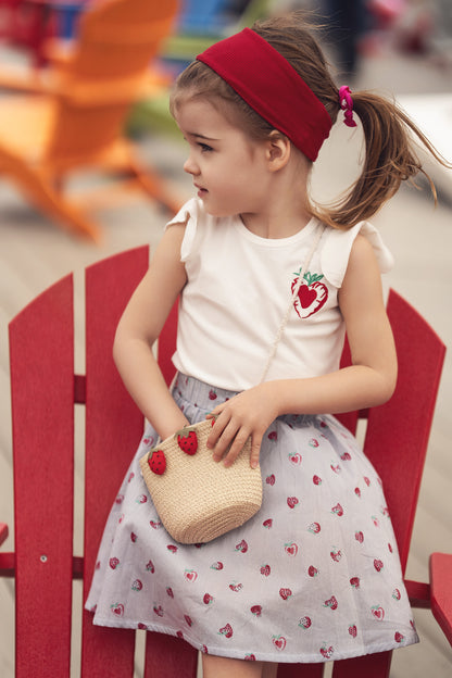 Young girl wearing a strawberry-print skirt with white top, red headband, and a straw bag with strawberries.