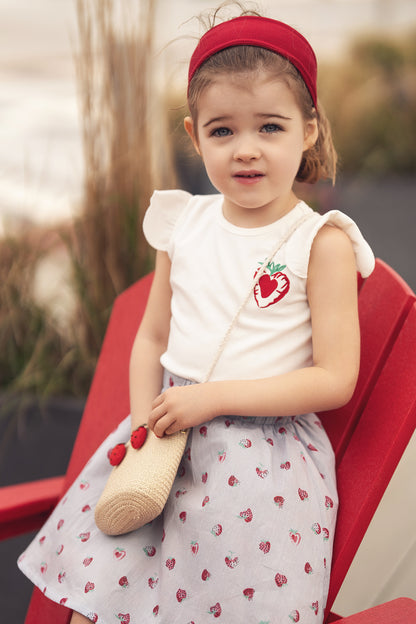 Young girl wearing a strawberry-print skirt with a white top, red headband, and straw bag.