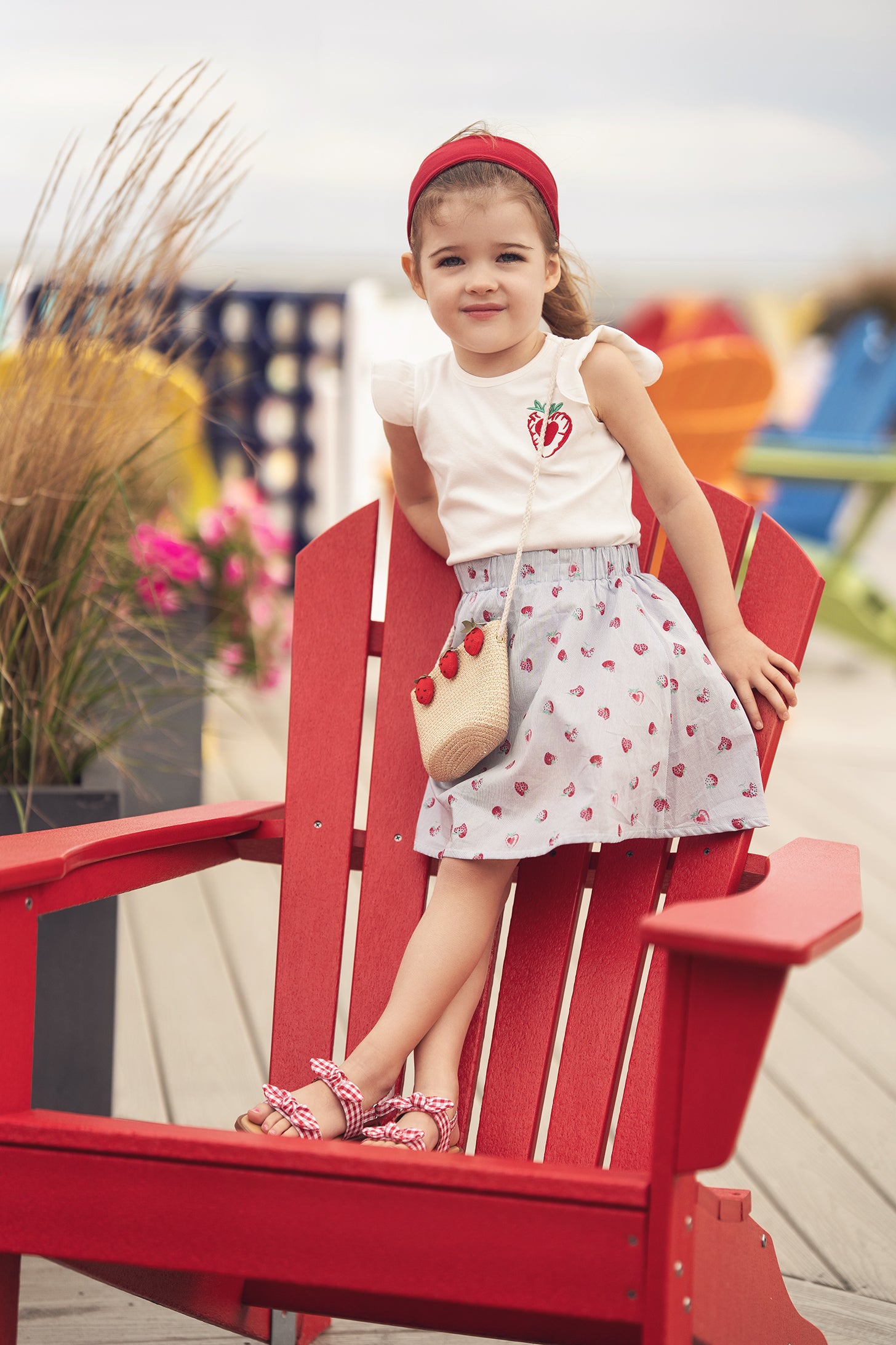 Girl wearing a strawberry-print skirt with white top and red headband, seated on a red Adirondack chair outdoors.
