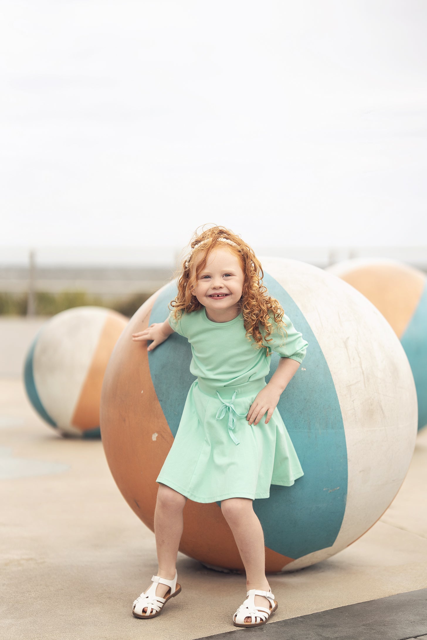 Young girl in mint green 3/4 puff sleeve T-shirt and skirt, smiling beside large striped playground balls.