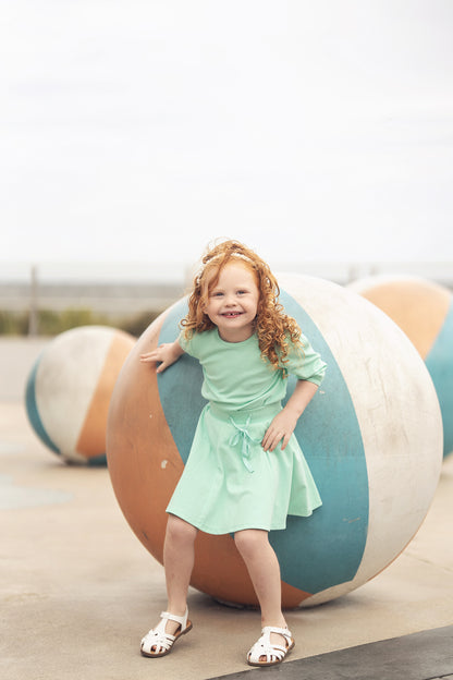 Young girl in mint green 3/4 puff sleeve T-shirt and skirt, smiling beside large striped playground balls.