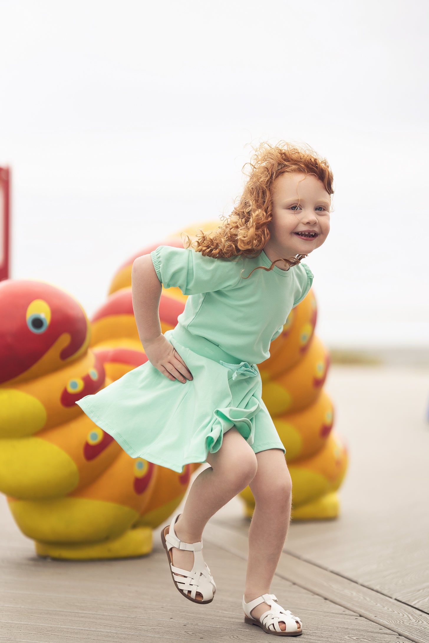 Girl wearing mint green 3/4 puff sleeve T-shirt, running on a boardwalk beside colorful sculptures.
