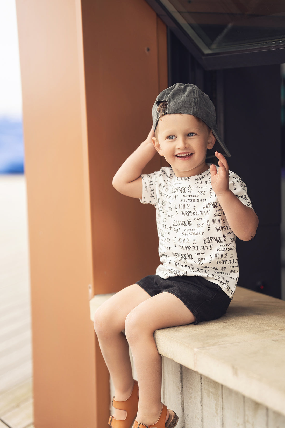 Happy toddler boy wearing a white Number Print T-shirt with coordinate-style black numbers and a backwards cap.