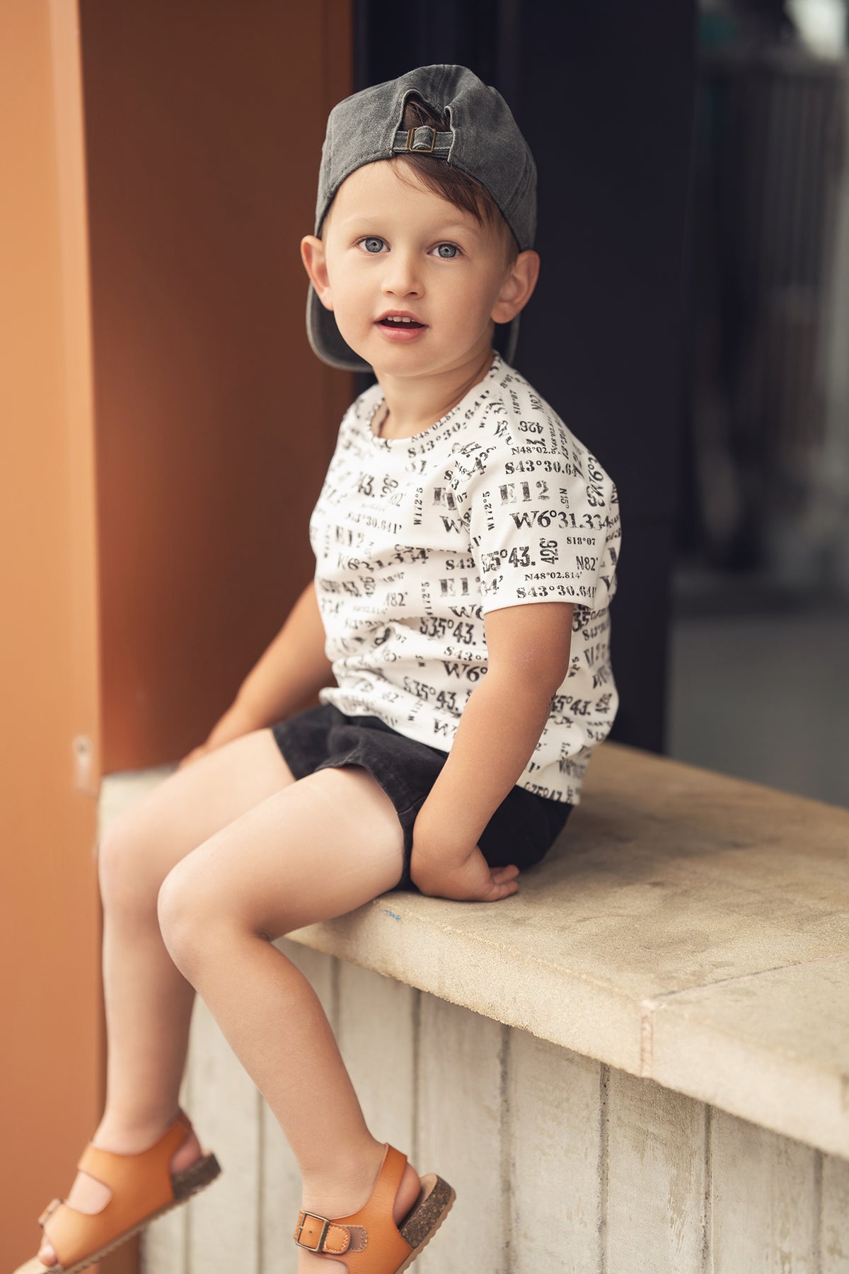Toddler boy in a white number-print T-shirt with coordinate numbers, black shorts, orange sandals, and a gray cap.