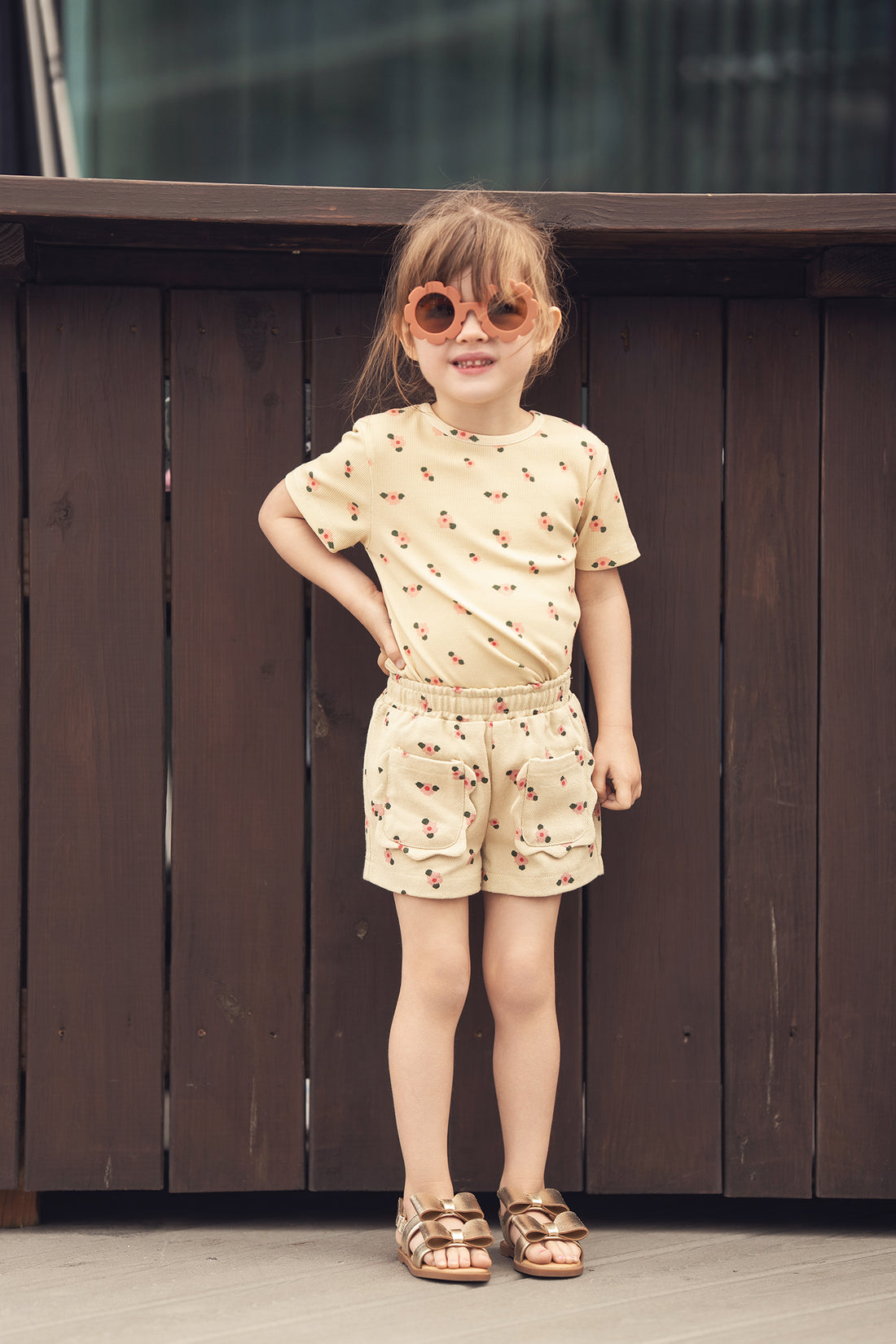 Tan Floral Shorts with pockets worn by a girl in a matching floral top, pink sandals, and flower-shaped sunglasses.