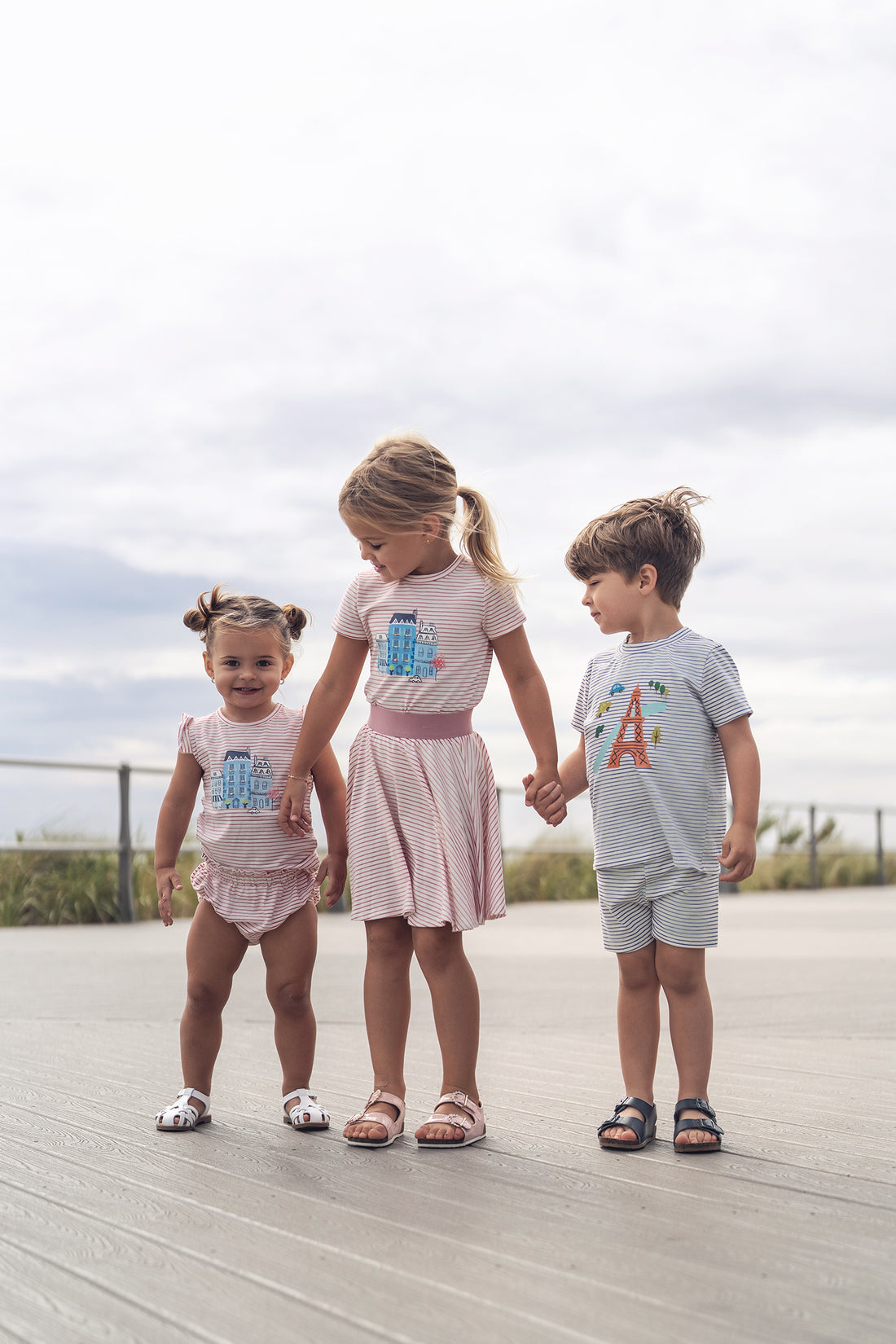 Three kids wear Eiffel Tower Shorts Set in pastel striped outfits on a boardwalk.