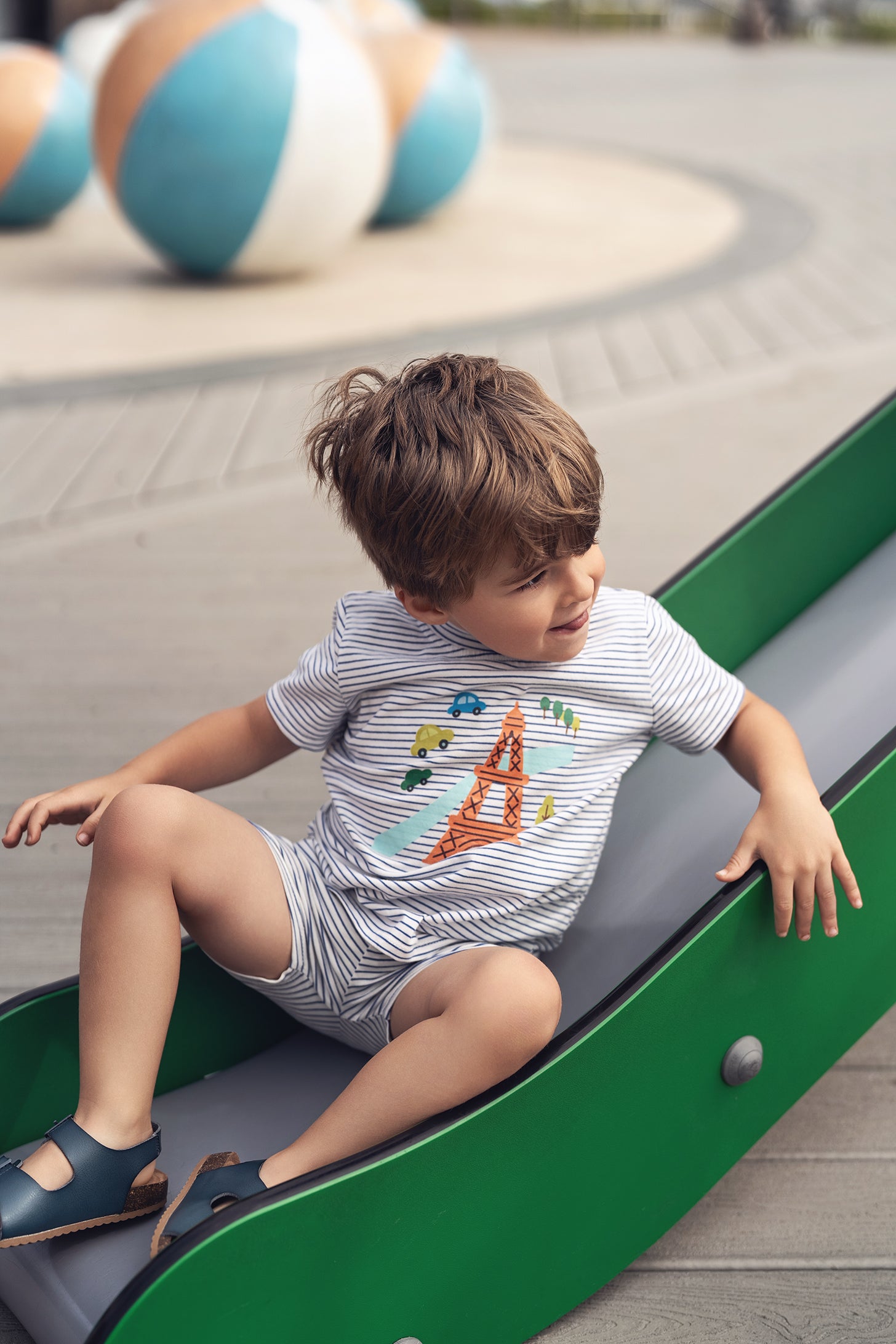 Child wearing Eiffel Tower Shorts Set on a green slide outdoors, with colorful balls in the background.
