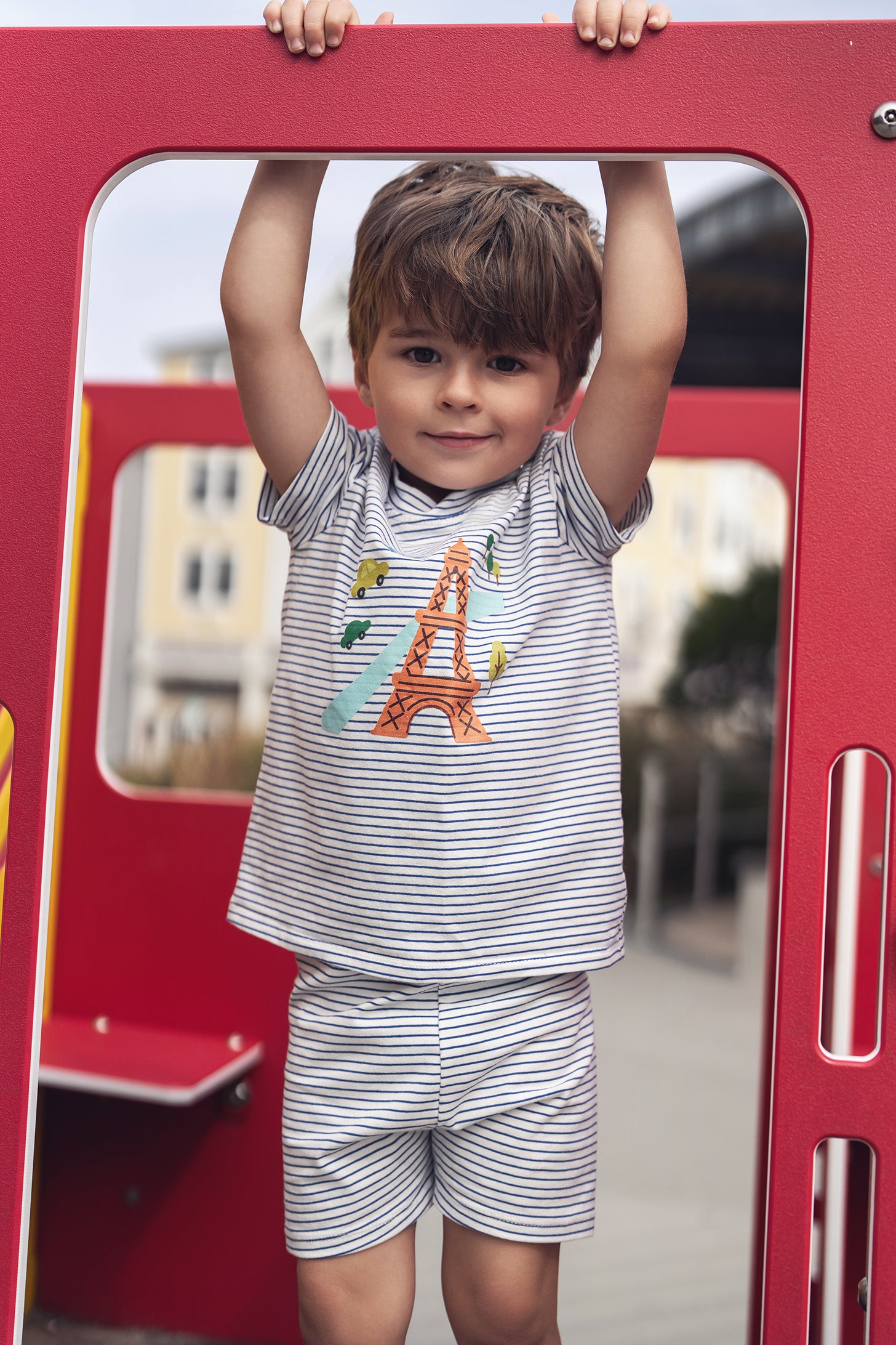 Child wearing Eiffel Tower Shorts Set, striped tee with Eiffel Tower graphic, hanging from a red playground frame.