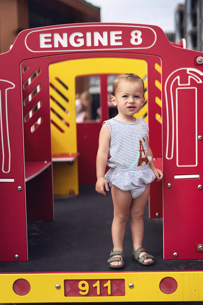 Toddler in Eiffel Tower bloomers &amp; striped tank set, standing at a colorful playground.