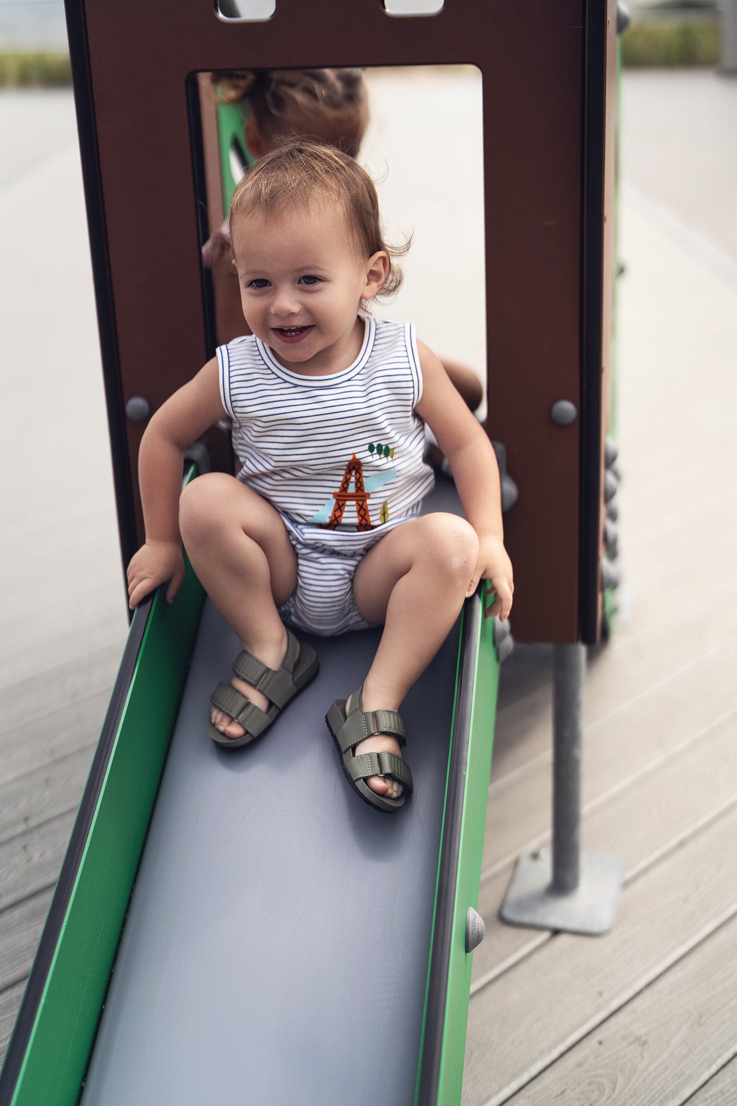 Toddler in striped tank top with Eiffel Tower graphic and bloomers on a playground slide, smiling.
