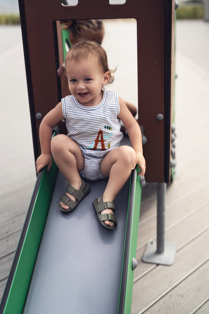 Toddler in striped tank top with Eiffel Tower graphic and bloomers on a playground slide, smiling.