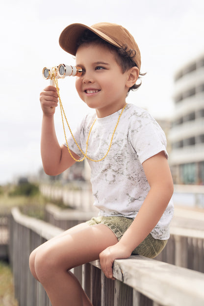 Smiling boy wearing Map T-shirt with vintage map print, tan cap, and binoculars on a boardwalk.