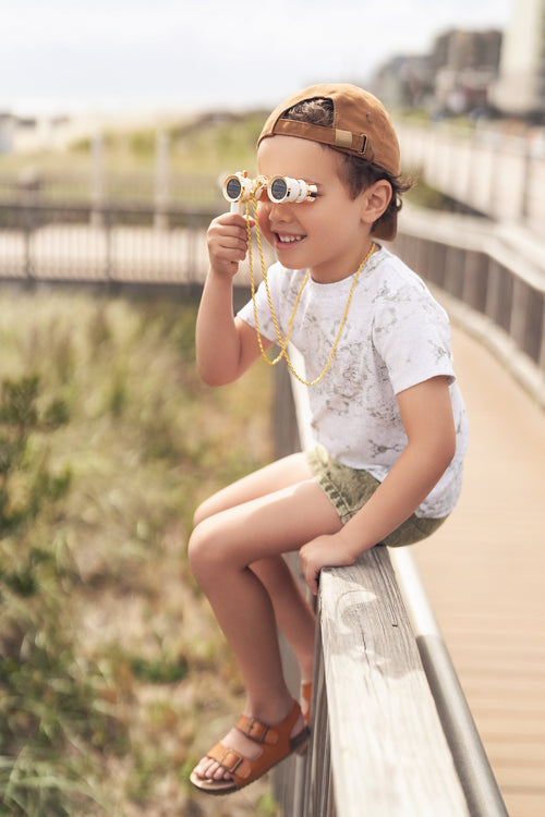Young boy in Map T-shirt with vintage map print, outdoors, wearing cap and binoculars.