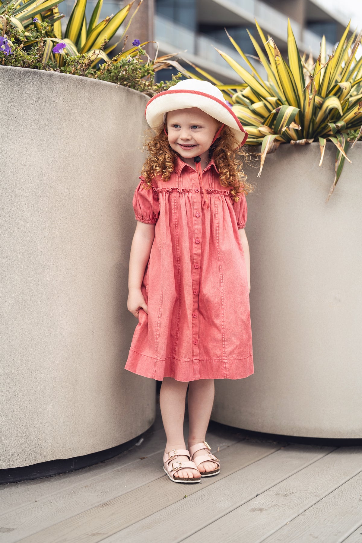 Little girl in Short Sleeve Dress - Pink Denim with button front, outdoors beside planters.