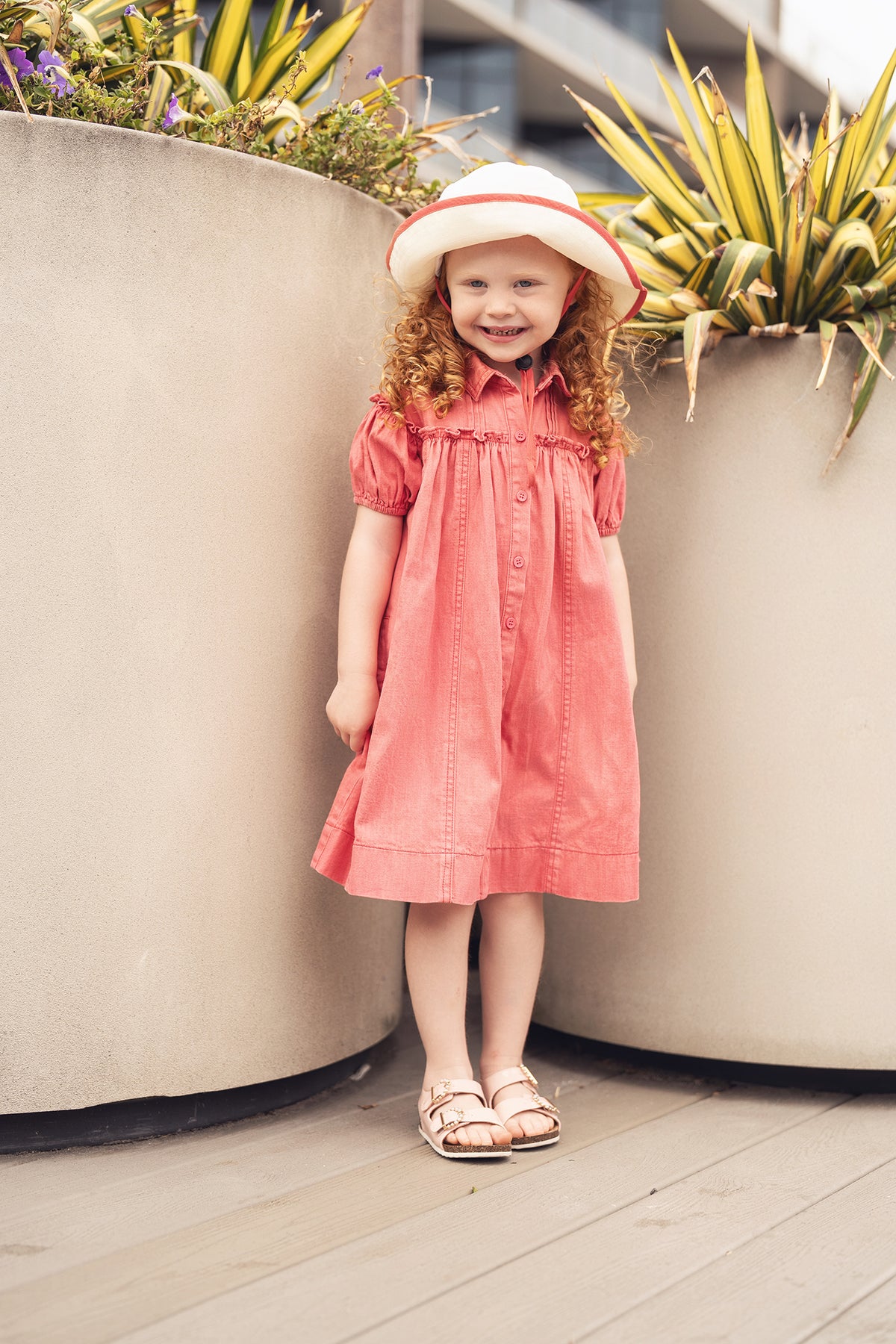 Young girl in a pink denim 3/4 sleeve dress, button front, with white hat, beside large planters.