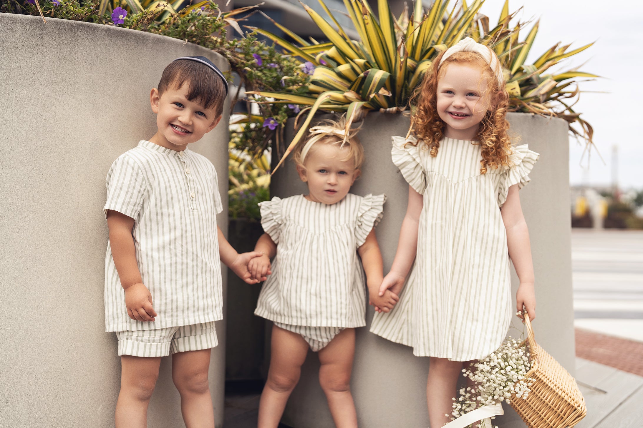 Three children in matching green-striped outfits pose outdoors beside planters.