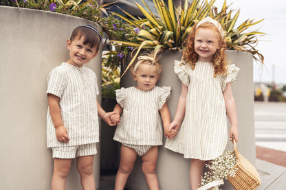 Three children in matching green-striped outfits pose outdoors beside planters.