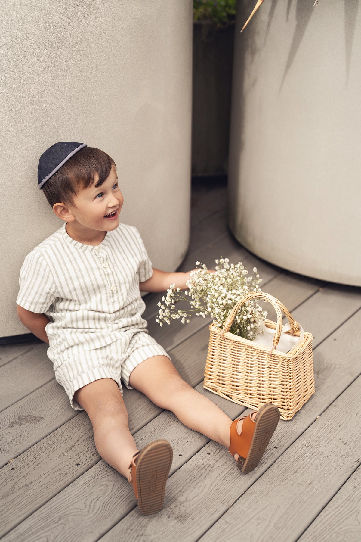 Young child in a green striped shirt sits on a wooden deck beside a woven basket of flowers.