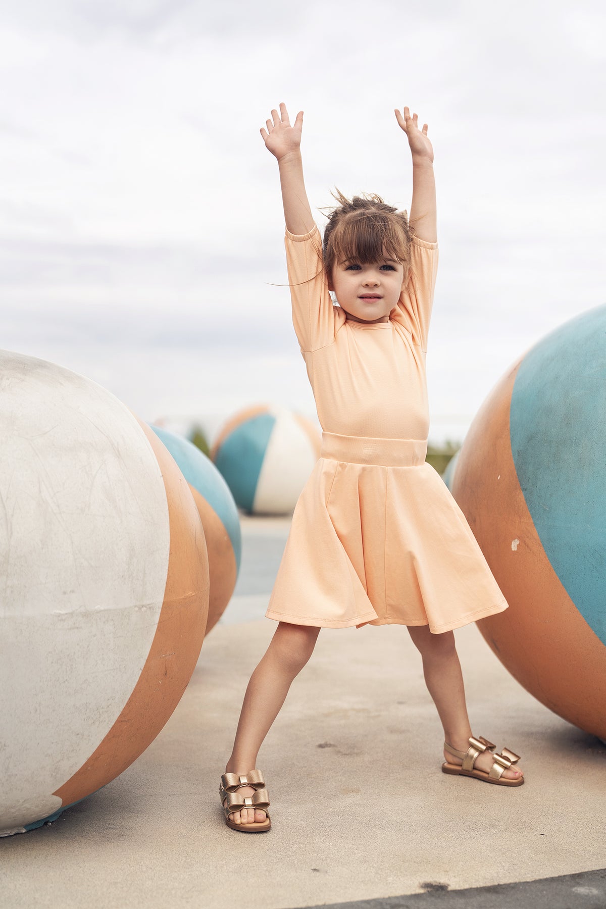 Peach 3/4 puff sleeve T-shirt with matching skirt worn by a girl, arms raised, outdoors among pastel spheres.