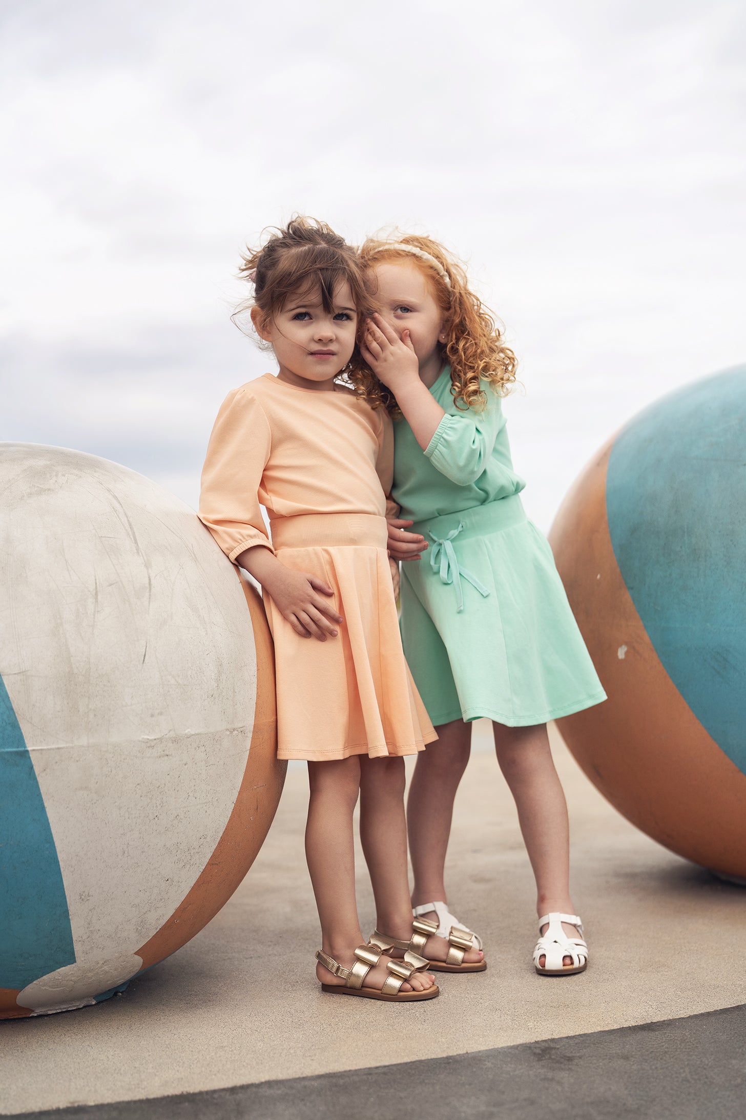 Two young girls in peach and mint green outfits pose beside large striped beach balls.