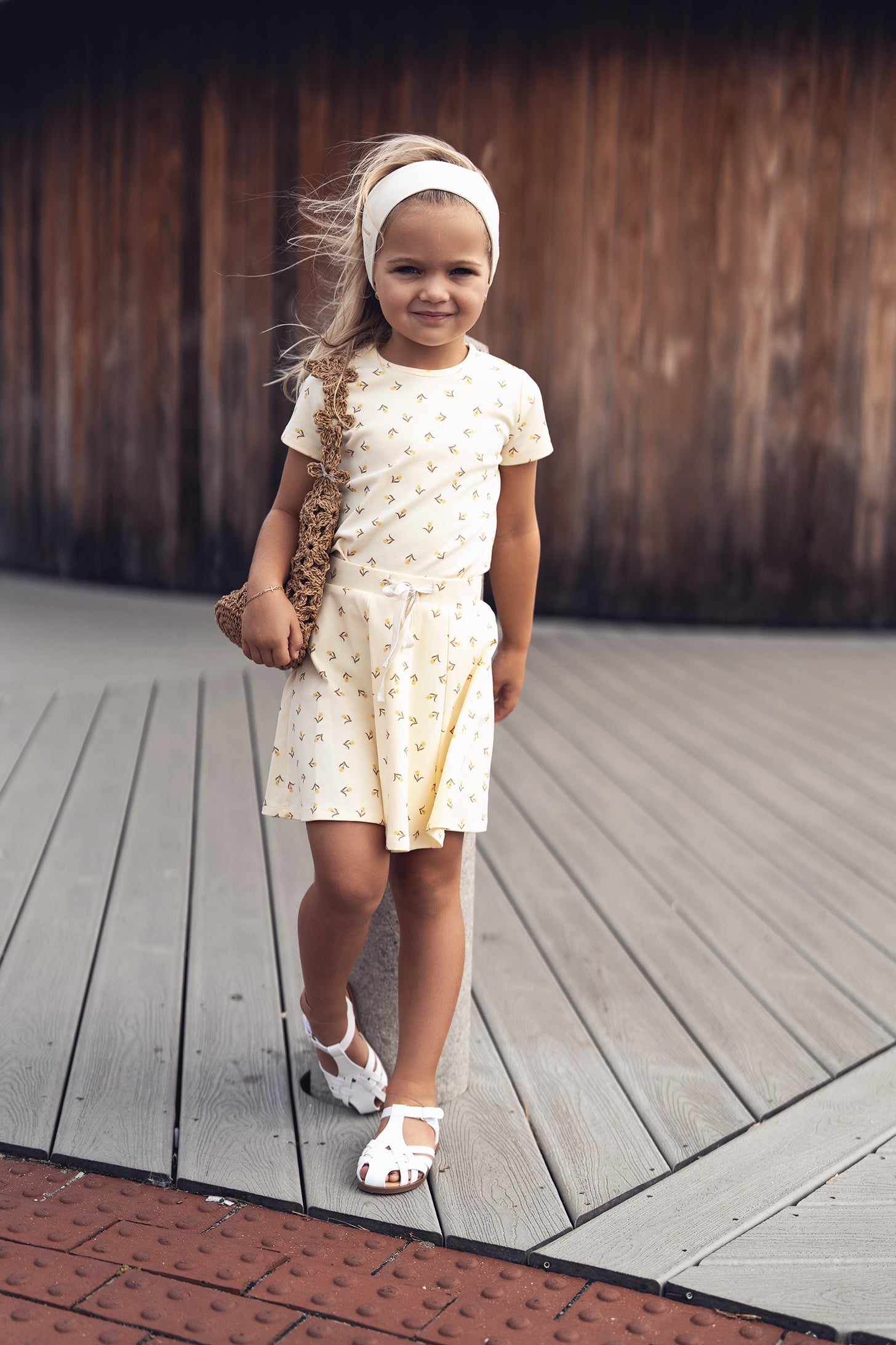 Young girl in a butter-yellow tulip-pattern short-sleeve outfit, white headband, sandals, and a woven bag.