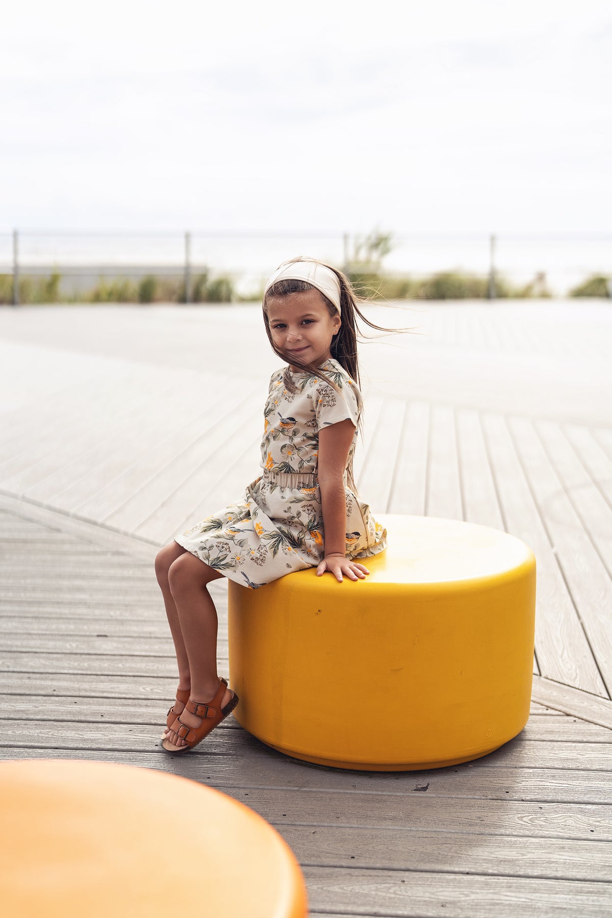 Girl wearing floral Garden Finch Circle Skirt sits on a yellow circular seat on a wooden boardwalk by the beach.