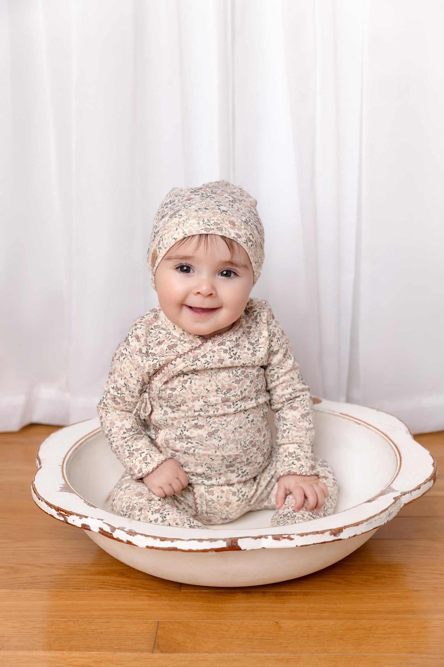 Purple Flower Stretchy girls stretchies by Adee, floral print baby outfit with hat, seated in a shallow wooden bowl.