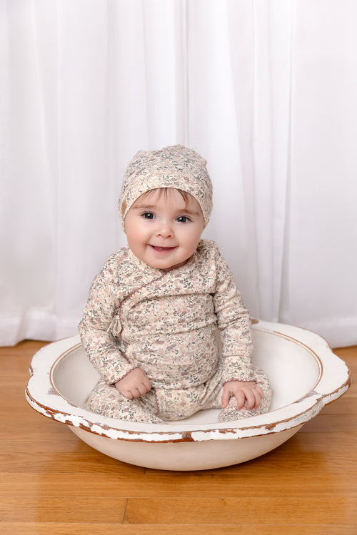 Purple Flower Stretchy girls stretchies by Adee, floral print baby outfit with hat, seated in a shallow wooden bowl.